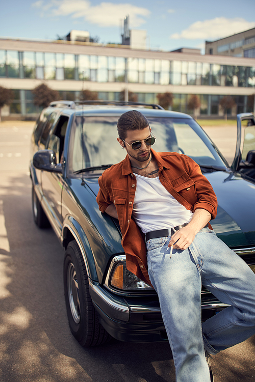 good looking sexy man in trendy bright attire posing next to his car with cigarette in his hand