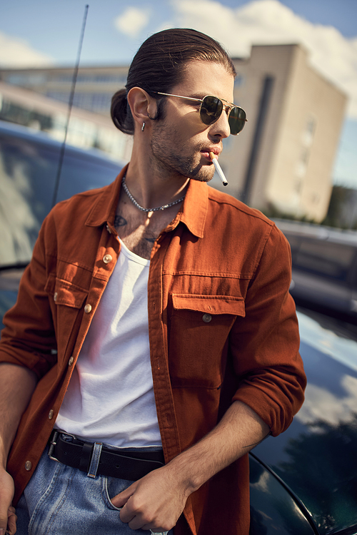 vertical shot of handsome young man in vibrant attire posing with cigarette next to his car