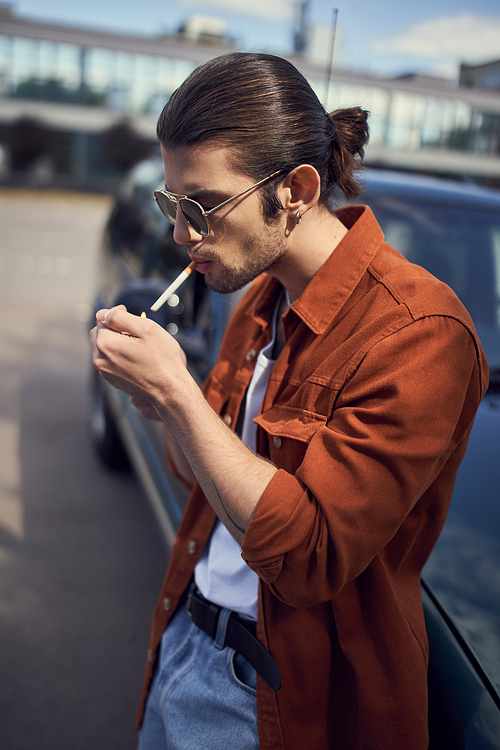 handsome young male model with ponytail and sunglasses in brown shirt lighting up his cigarette