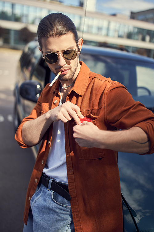 appealing young man with ponytail and sunglasses posing next to his car with cigarette in his mouth