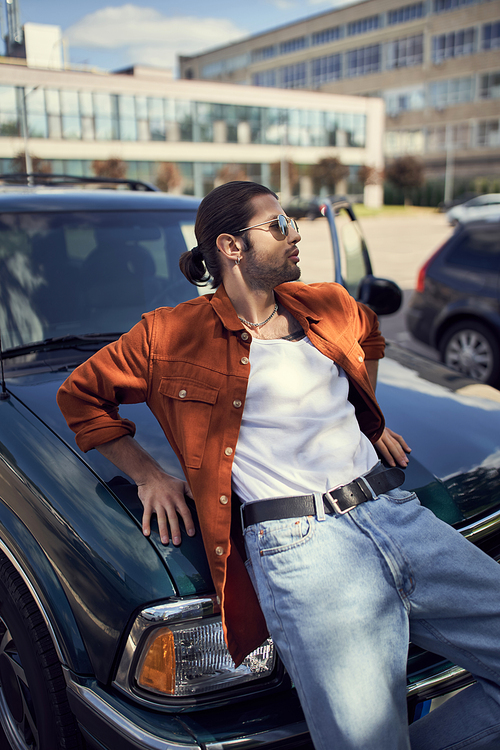 handsome young man with sunglasses and ponytail in vivid attire leaning on his car, fashion concept