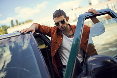 handsome sexy man with sunglasses and ponytail in vibrant outfit posing next to his car