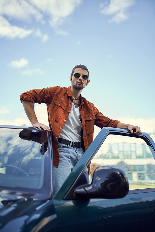 vertical shot of appealing young man in stylish vibrant clothing posing next to his car, fashion