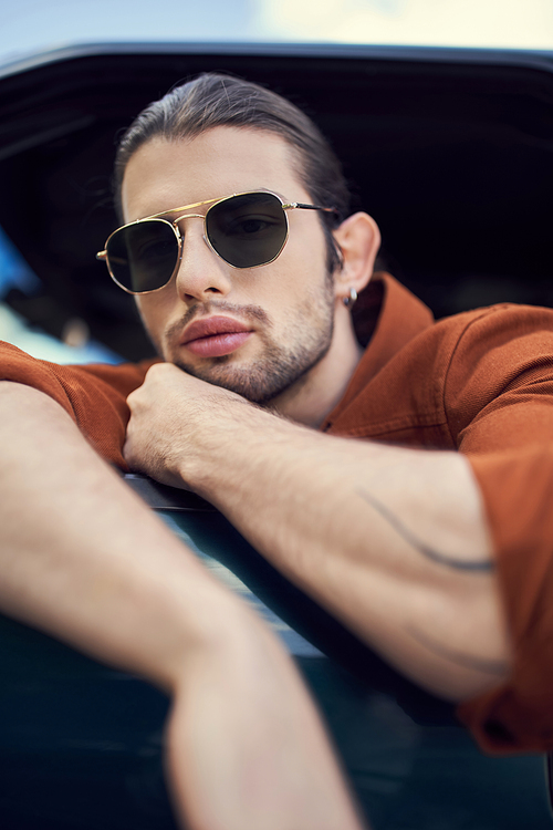 good looking stylish male model in sunglasses posing in his car with crossed arms, hand near face