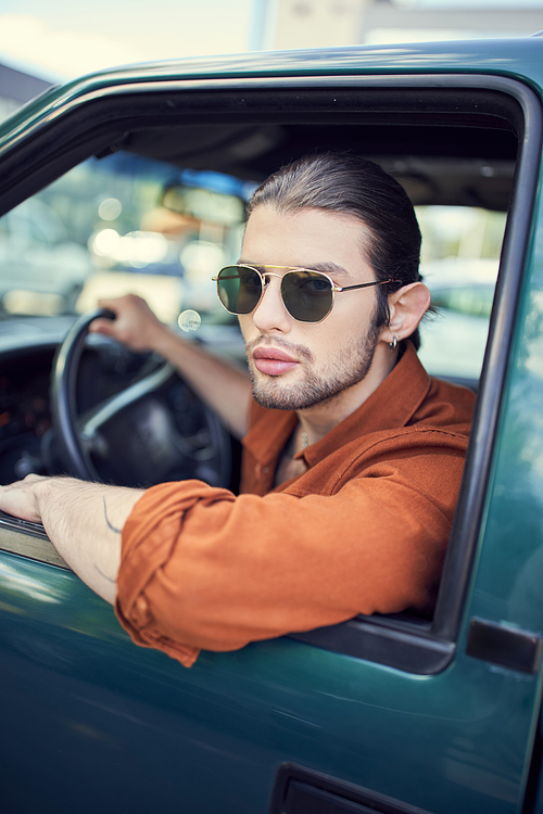 appealing young man with earring in trendy brown shirt with sunglasses looking out of car window