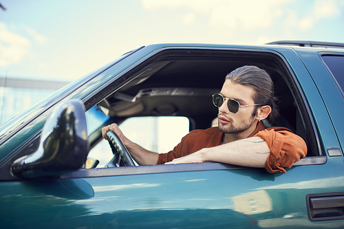 appealing young man with ponytail and earring in brown shirt looking out of car window, sexy driver