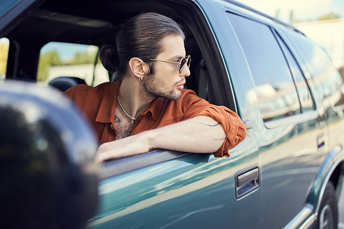 handsome man in stylish vivid outfit with sunglasses and ponytail looking out of car window, style