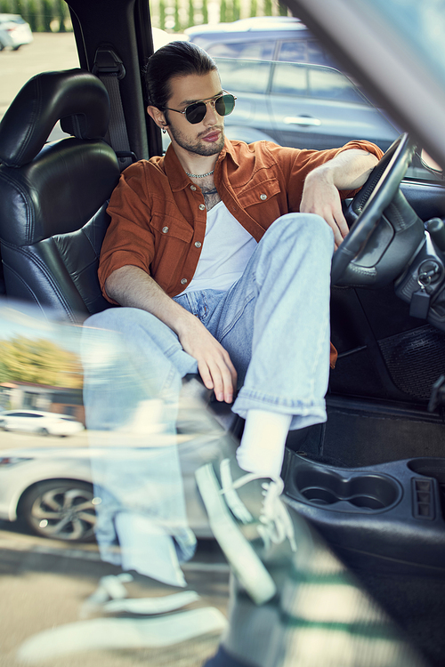 relaxed appealing man with sunglasses in jeans and brown shirt chilling in his car, fashion concept