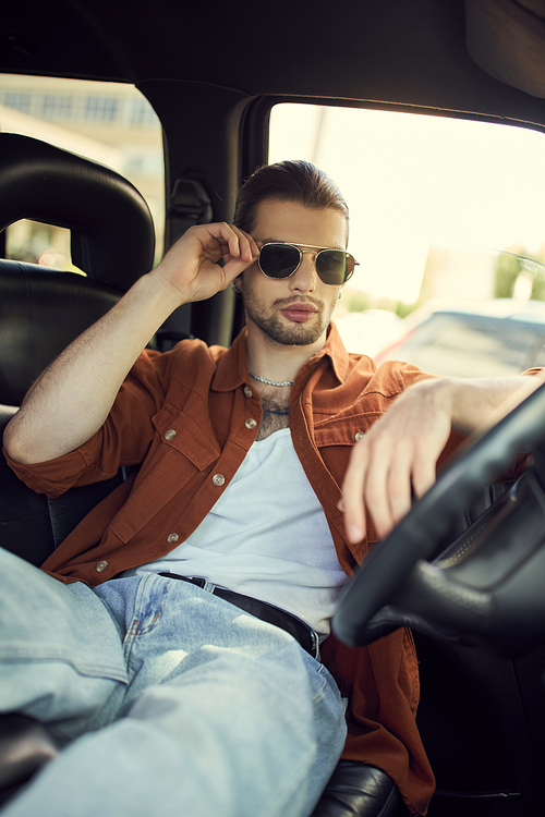 good looking young man in stylish outfit touching his sunglasses while relaxing in car, sexy driver
