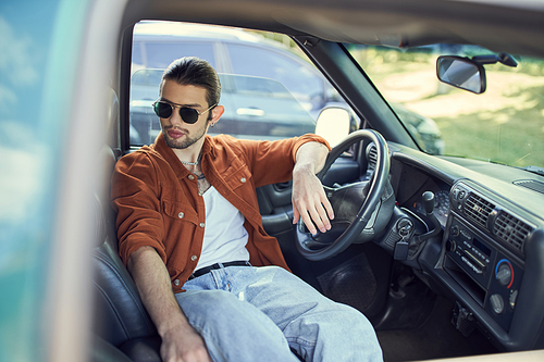 handsome man in trendy brown shirt and jeans chilling in his car behind steering wheel, fashion