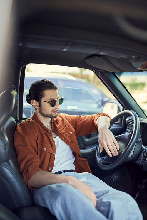charming man with ponytail and sunglasses relaxing behind steering wheel and looking away, fashion
