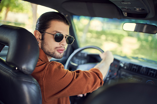 appealing stylish man with ponytail and sunglasses posing in his car and looking at camera