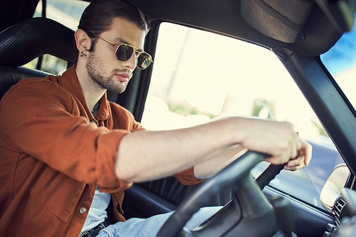 charming stylish male model in sunglasses with ponytail behind steering wheel of his car, style