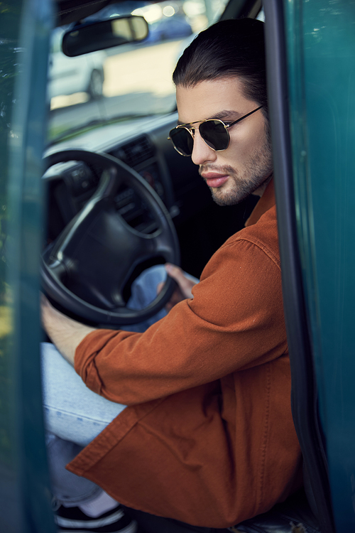 eye catching stylish man with sunglasses in brown shirt in his car with hands on steering wheel