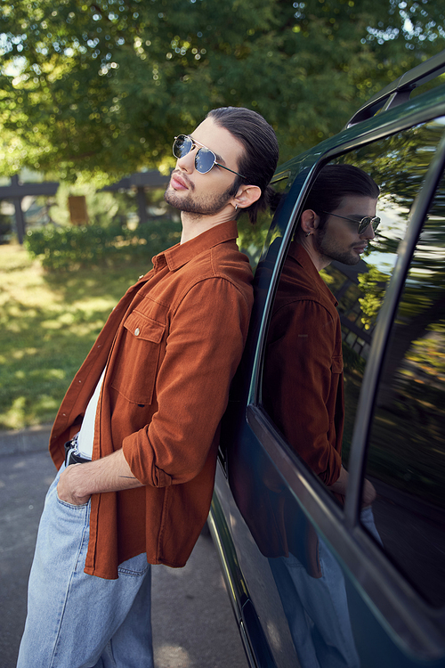 tempting stylish man with sunglasses posing outdoors leaning slightly on his car, fashion concept