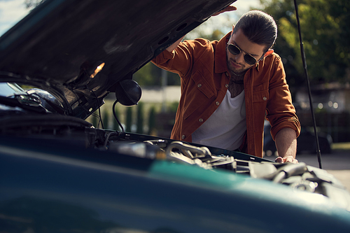 pensive stylish male model with ponytail and trendy accessories looking at engine of his car