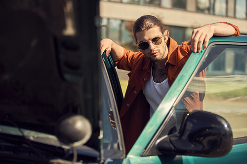 handsome sexy driver in brown shirt with sunglasses looking at camera posing next to his car