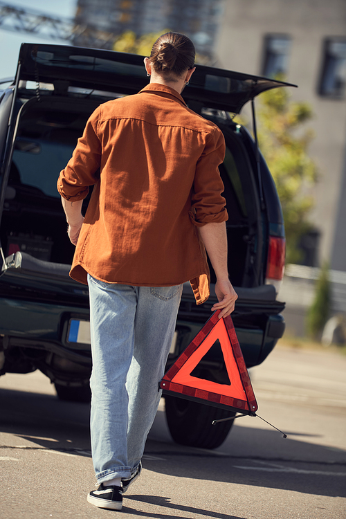 back view of young stylish man in urban attire walking to his car with warning triangle in hands