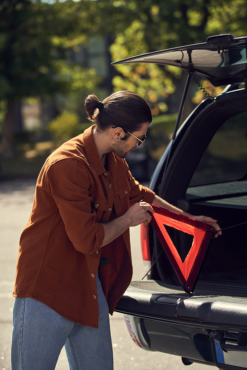 vertical shot of stylish man with sunglasses putting warning triangle in his car trunk, sexy driver