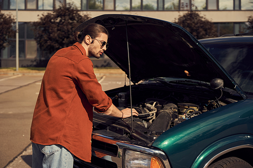 handsome bearded man with ponytail and sunglasses standing next to his car with opened engine hood