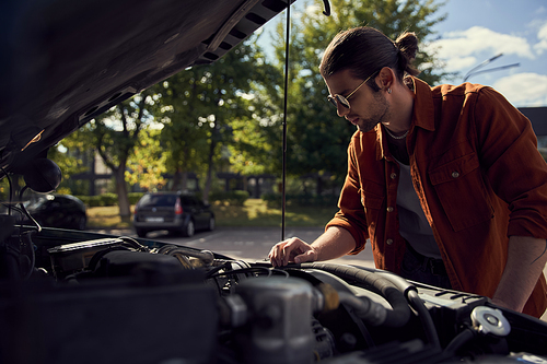 pensive handsome man in stylish outfit with sunglasses and ponytail checking on his car engine