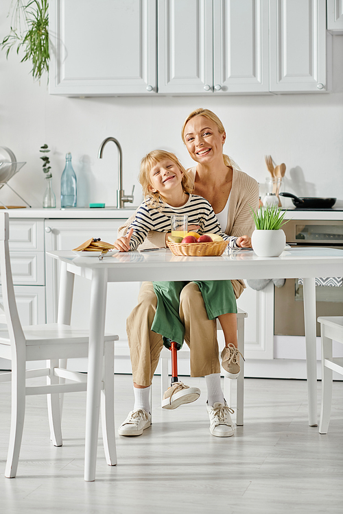 cheerful girl with prosthetic leg sitting on laps of happy mother during breakfast in kitchen