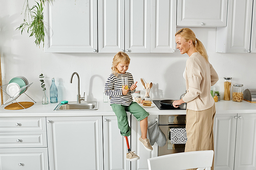 happy girl with prosthetic leg sitting on countertop with orange juice near mother cooking pancakes