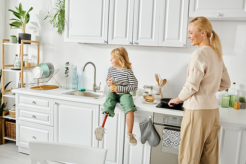 happy girl with prosthetic leg sitting on countertop with orange juice near mother cooking pancakes