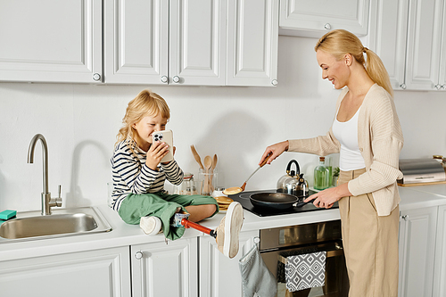 happy girl with prosthetic leg sitting on countertop with smartphone near mother cooking pancakes