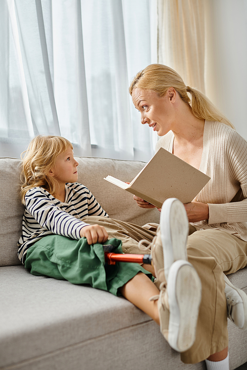 blonde mother reading book to cute daughter with prosthetic leg and sitting together in living room