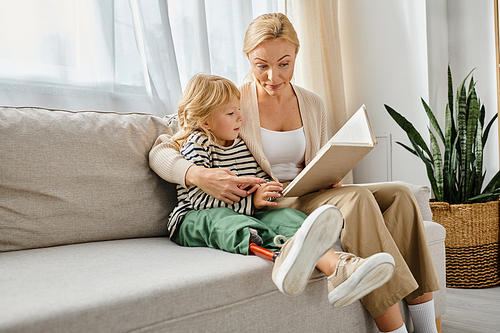 blonde woman reading book to daughter with prosthetic leg while sitting together in living room