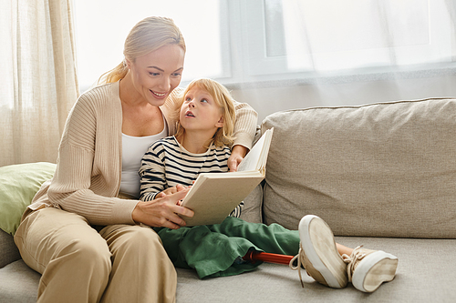 happy mother reading book to her daughter with prosthetic leg and sitting together in living room