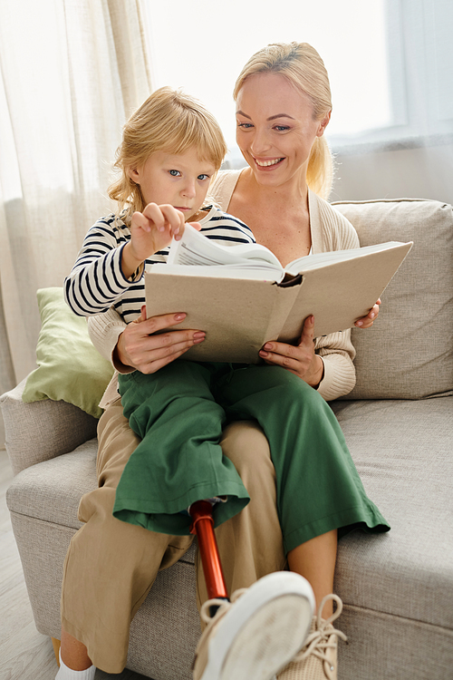 happy girl with prosthetic leg sitting on laps of her blonde mother and reading book in living room