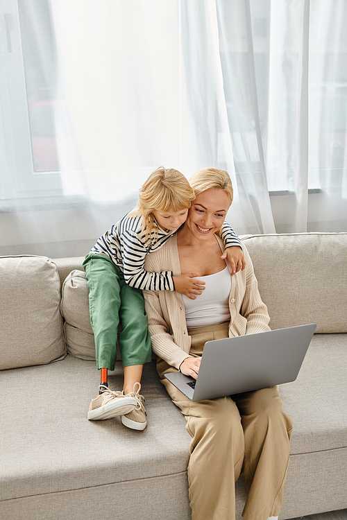 happy child with prosthetic leg hugging blonde mother working on laptop in modern living room