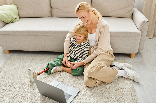 happy child with prosthetic leg watching movie on laptop while sitting on carpet with mother