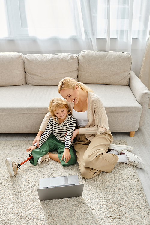 happy child with prosthetic leg watching movie on laptop and sitting on carpet together with mother