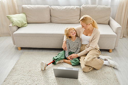 happy girl with prosthetic leg watching movie on laptop and sitting on carpet together with mother