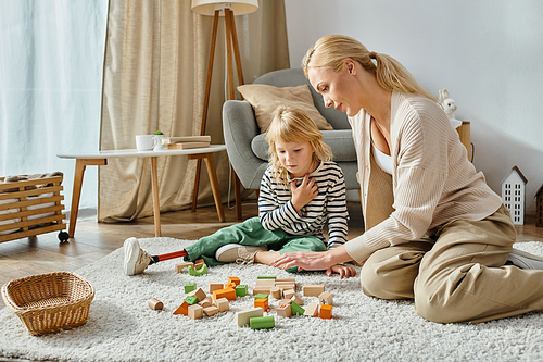 disabled girl with prosthetic leg sitting on carpet and looking at wooden toys near mother