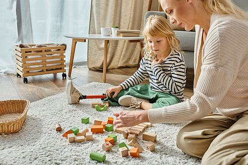 disabled little girl with prosthetic leg sitting on carpet and looking at wooden toys near mother