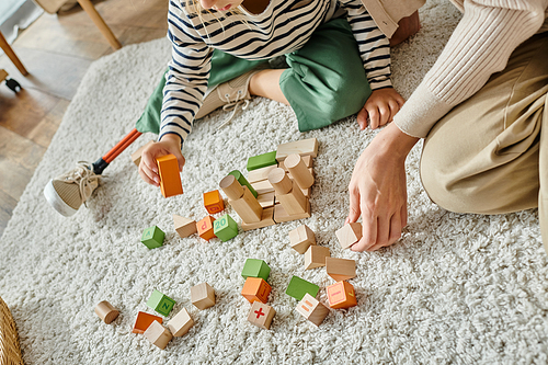 cropped girl with prosthetic leg sitting on carpet and playing with wooden toys near mother