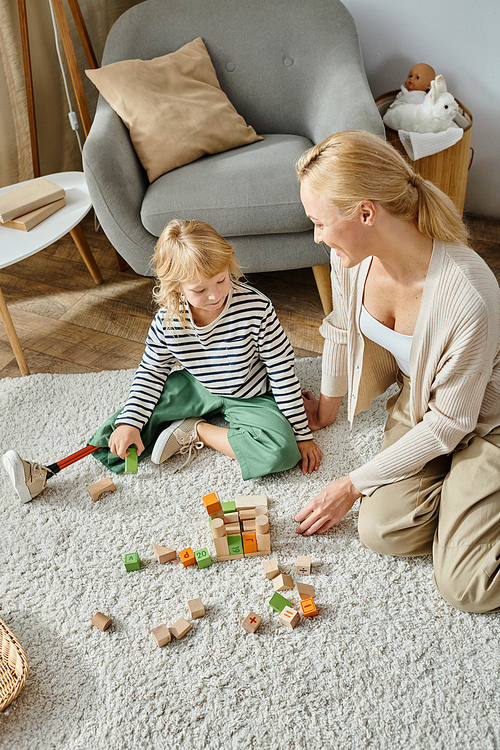 little girl with prosthetic leg sitting on carpet and playing with wooden toys near happy mother