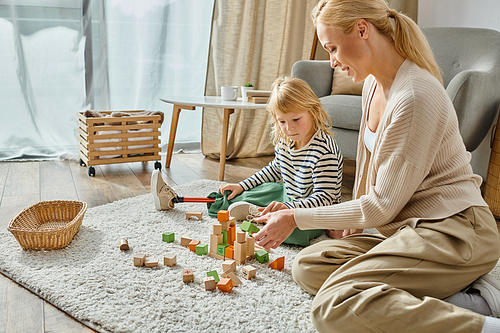 little girl with prosthetic leg sitting on carpet and playing with wooden toys near joyful mother