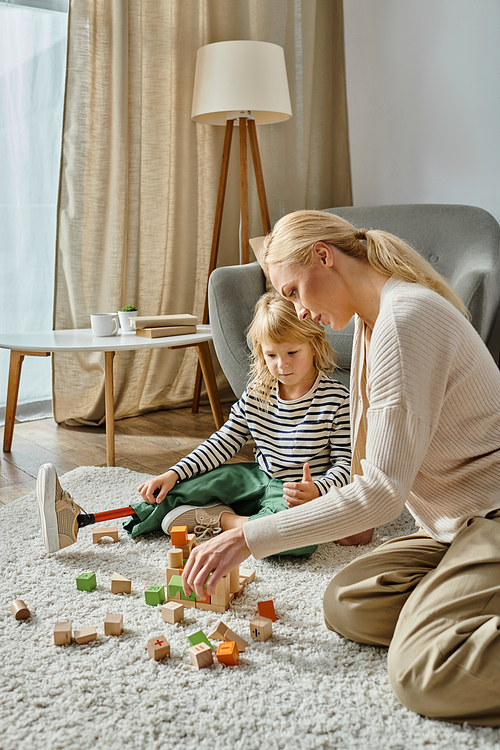 disabled girl with prosthetic leg sitting on carpet and playing with wooden toys near blonde mother