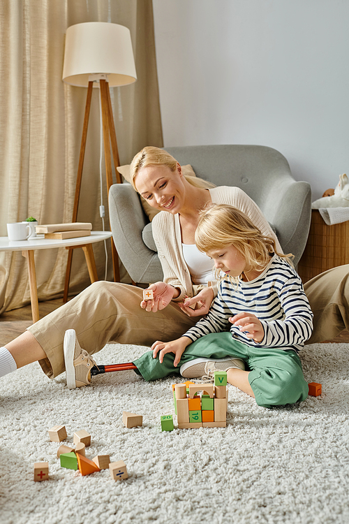 little girl with prosthetic leg sitting on carpet and playing with wooden toys near cheerful mother