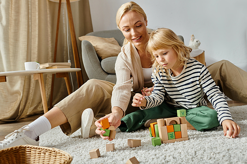 cure girl with prosthetic leg sitting on carpet and playing with wooden blocks near happy mother