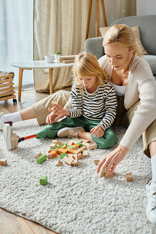 surprised girl with prosthetic leg sitting on carpet and playing with wooden blocks near mother