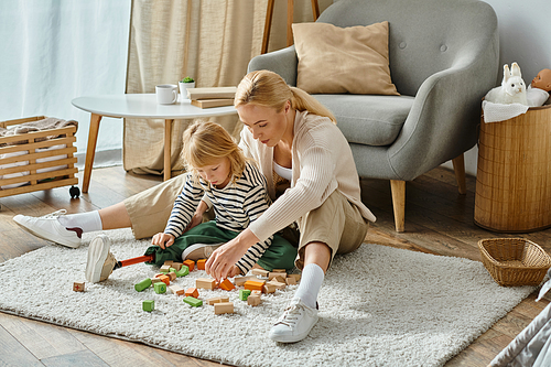 girl with prosthetic leg sitting on carpet and playing wooden blocks game near blonde mother