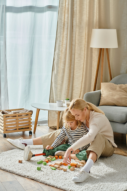 blonde girl with prosthetic leg sitting on carpet and playing wooden blocks game near mother