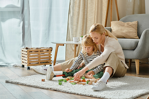 blonde girl with prosthetic leg sitting on carpet and playing wooden blocks game near caring mother