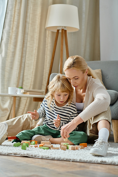 blonde girl with prosthetic leg sitting on carpet and playing with wooden blocks near caring mom